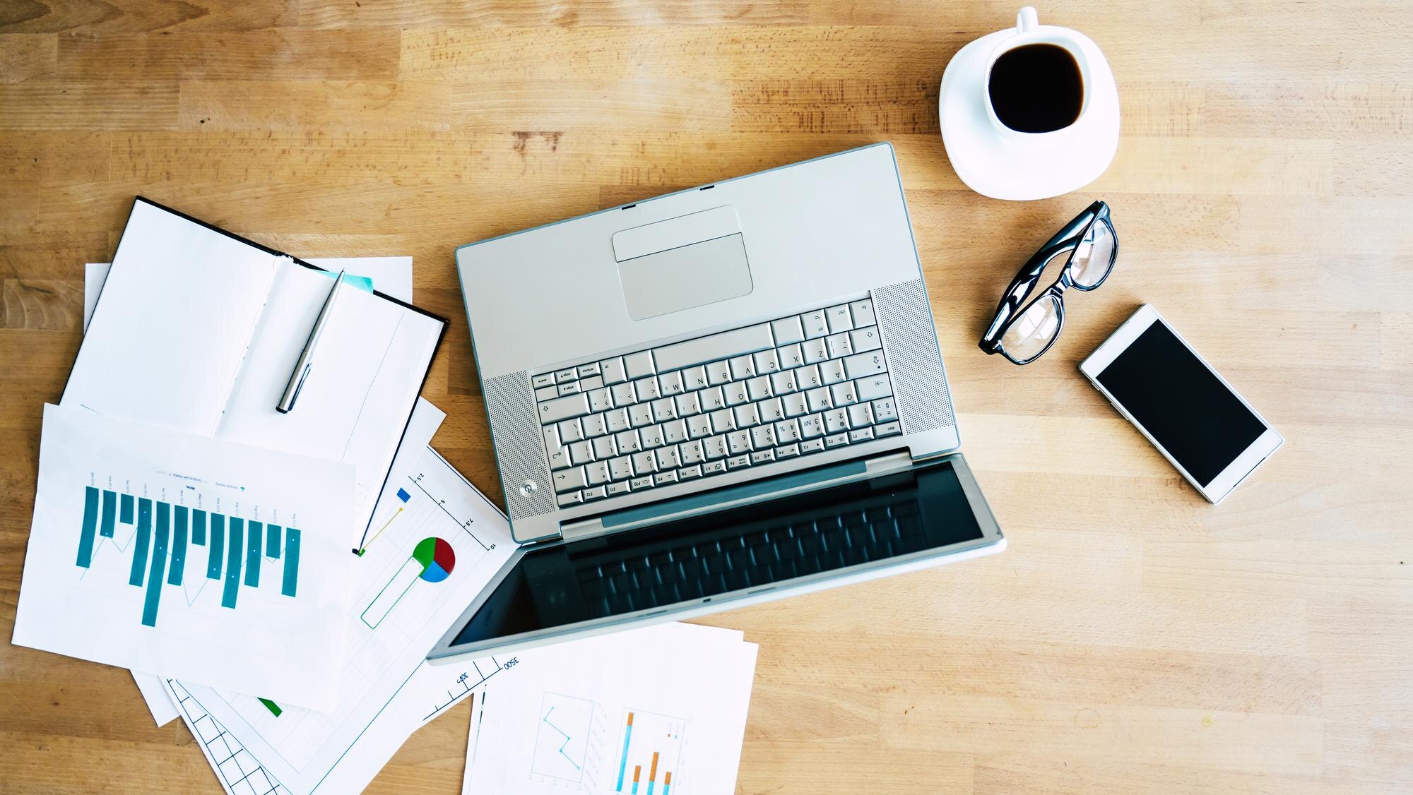 Overhead view of a laptop, printed reports and reading glasses on a wooden desk